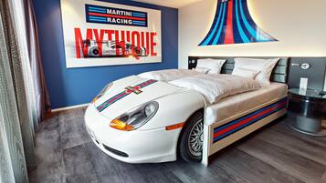 A bedroom with a bed featuring a racing car design, a wall with a racing theme, and a modern lamp.