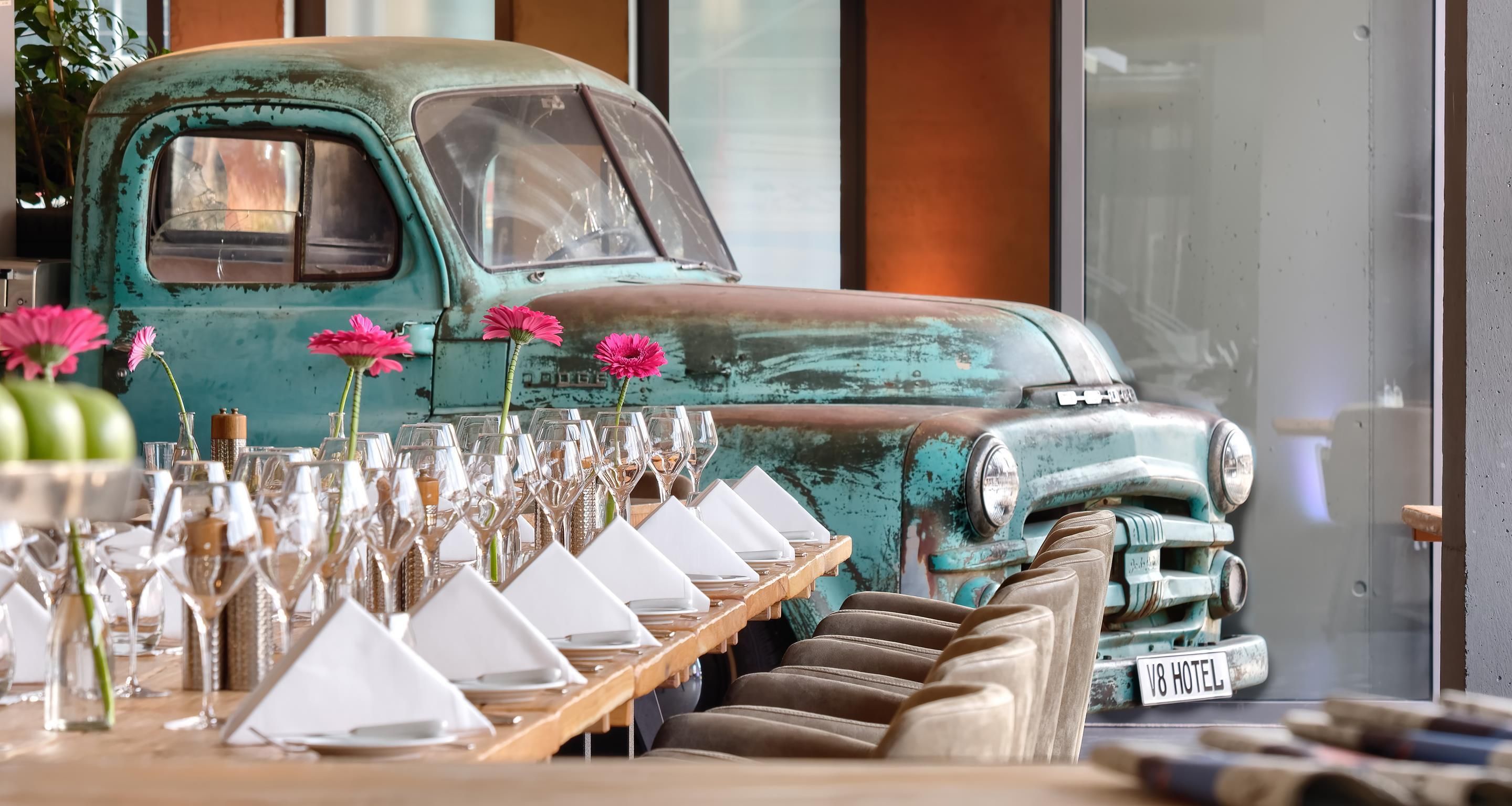 A vintage teal truck with a white license plate, parked in front of a table set with white napkins and pink flowers.