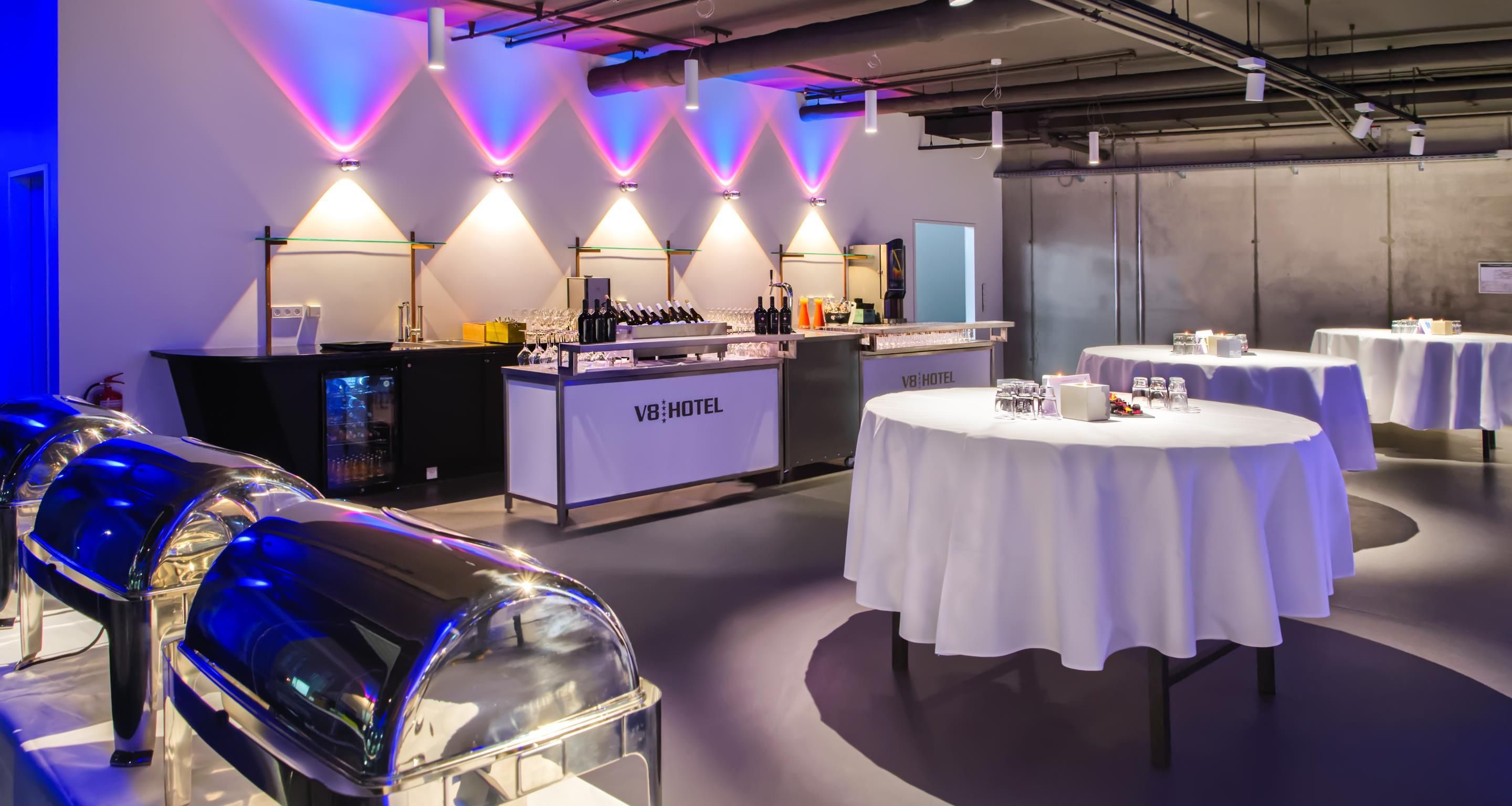 En moderne hotelbar med en bardisk fyldt med flasker og glas, omgivet af borde med hvide duge.
