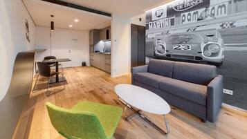 Ein modernes Wohnzimmer mit einem grauen Sofa, einem weißen Couchtisch und einem grünen Sessel. Im Hintergrund ist eine Küche erkennbar.
