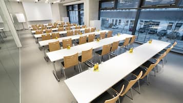 A conference room with multiple tables and chairs, large windows with a view of parked cars and buildings outside.