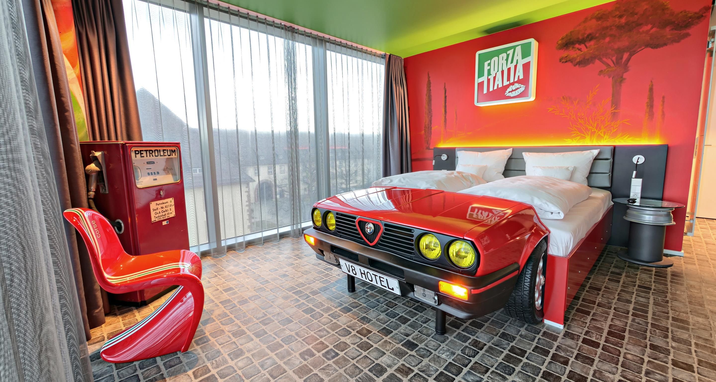 A hotel room with a bed, a red car-shaped headboard, a red chair, a wall with a 'Forza Italia' sign, and a window with curtains.