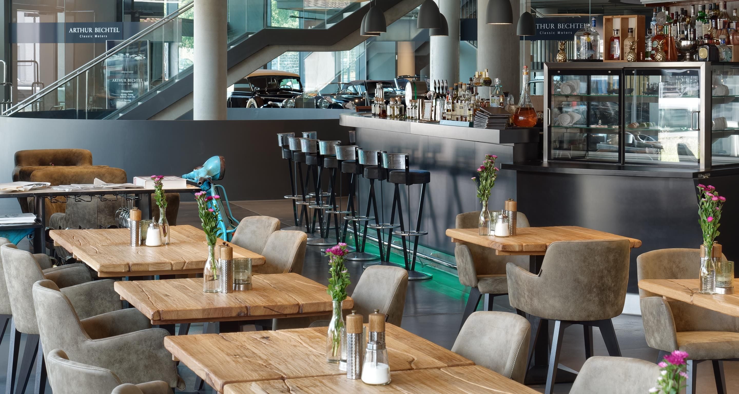 En modern restaurang med träbord och stolar, en bardisk med flaskor och utsikt över klassiska bilar genom stora fönster.