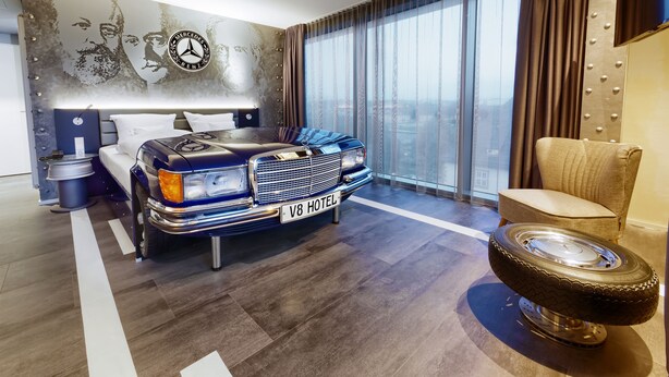 A modern hotel room with a classic car as a centerpiece, a large bed, a seating area, and a city view. - V8 Hotel Motorworld Region Stuttgart, BW Premier Collection (Boeblingen)