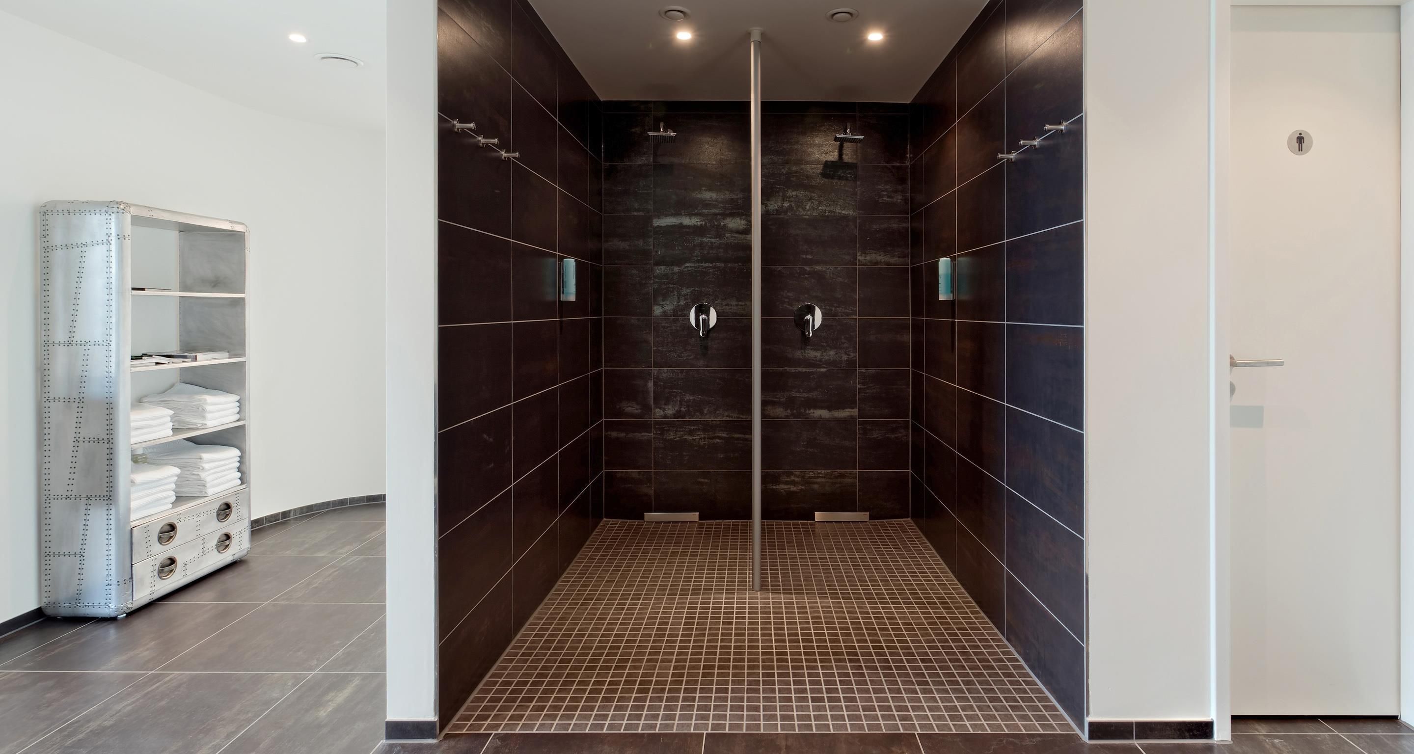 A modern bathroom with dark tiled walls, a shower area, and a storage unit with folded towels.