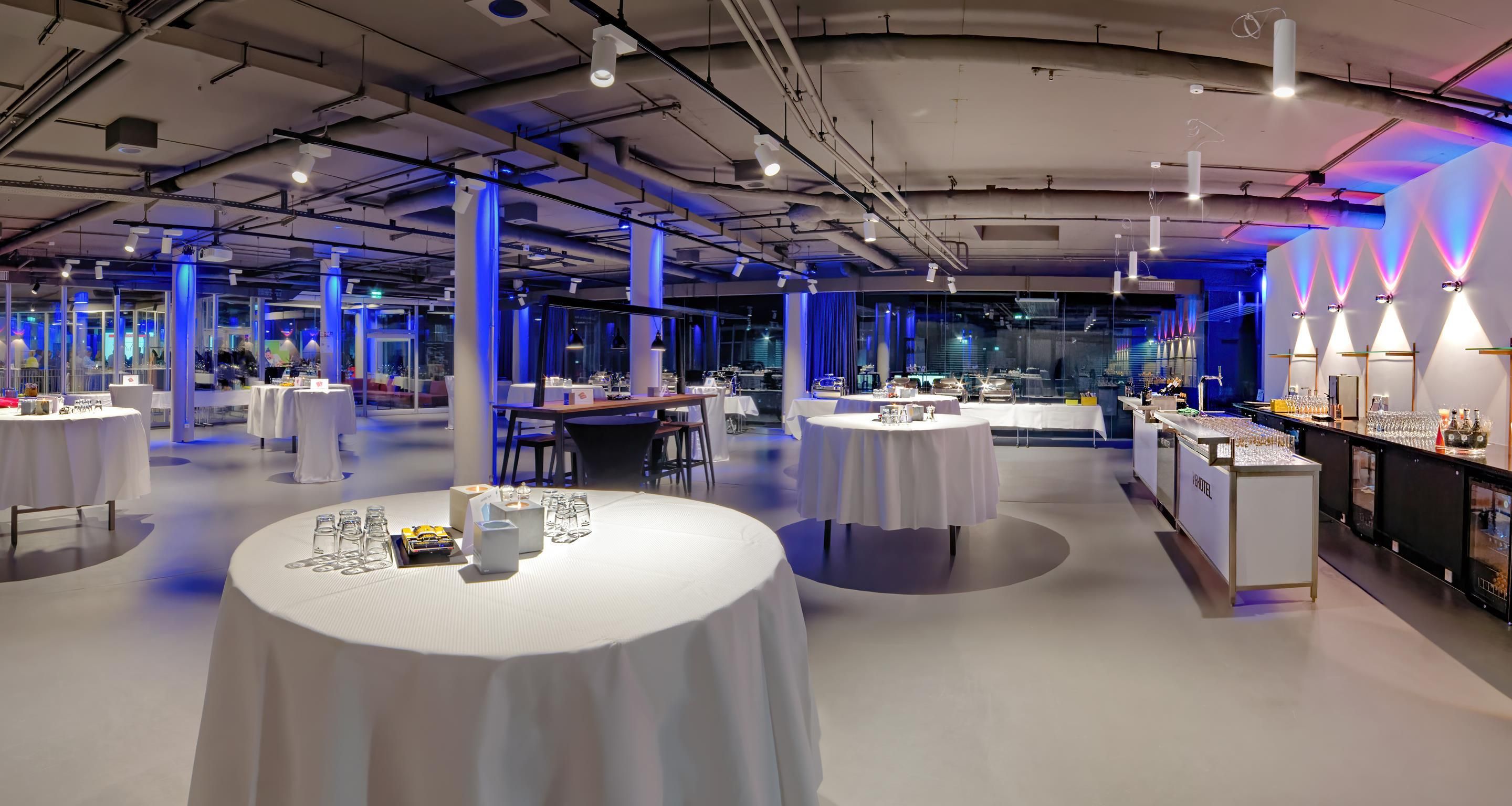 A modern hotel lobby with round tables, a bar area, and a view of the city through large windows.