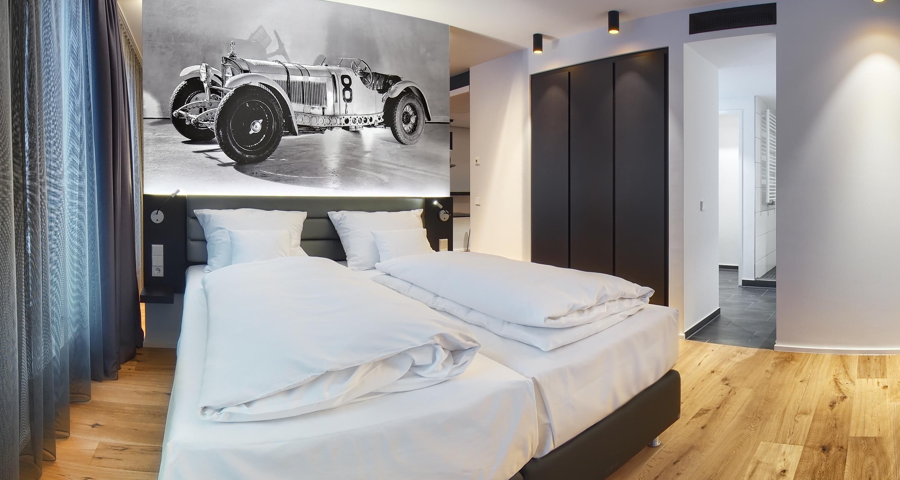 A modern bedroom with a large bed, a headboard with integrated lighting, a wall-mounted artwork of a vintage car, and a walk-in closet.