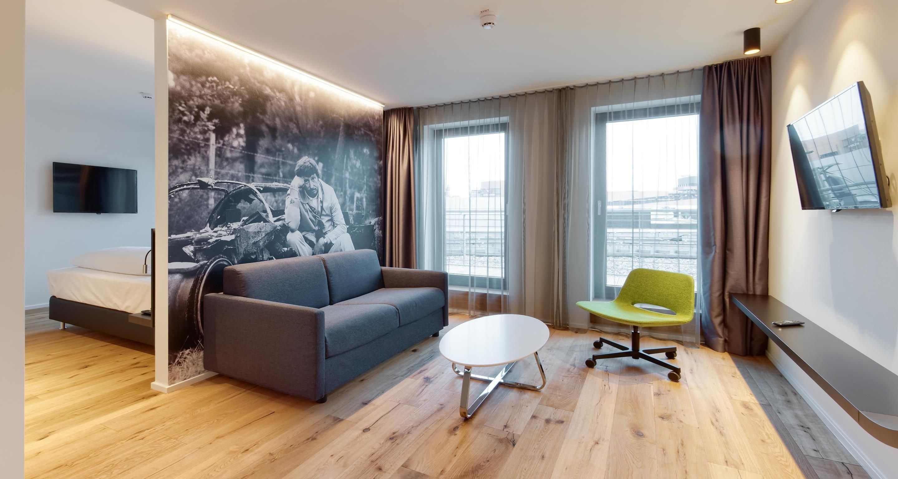 Ein modernes Hotelzimmer mit einer Couch, einem kleinen runden Tisch, einem grünen Sessel, einem Flachbildfernseher und einem großen Fenster mit Blick auf die Stadt.