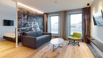 Ein modernes Hotelzimmer mit einer Couch, einem kleinen runden Tisch, einem grünen Sessel, einem Flachbildfernseher und einem großen Fenster mit Blick auf die Stadt.