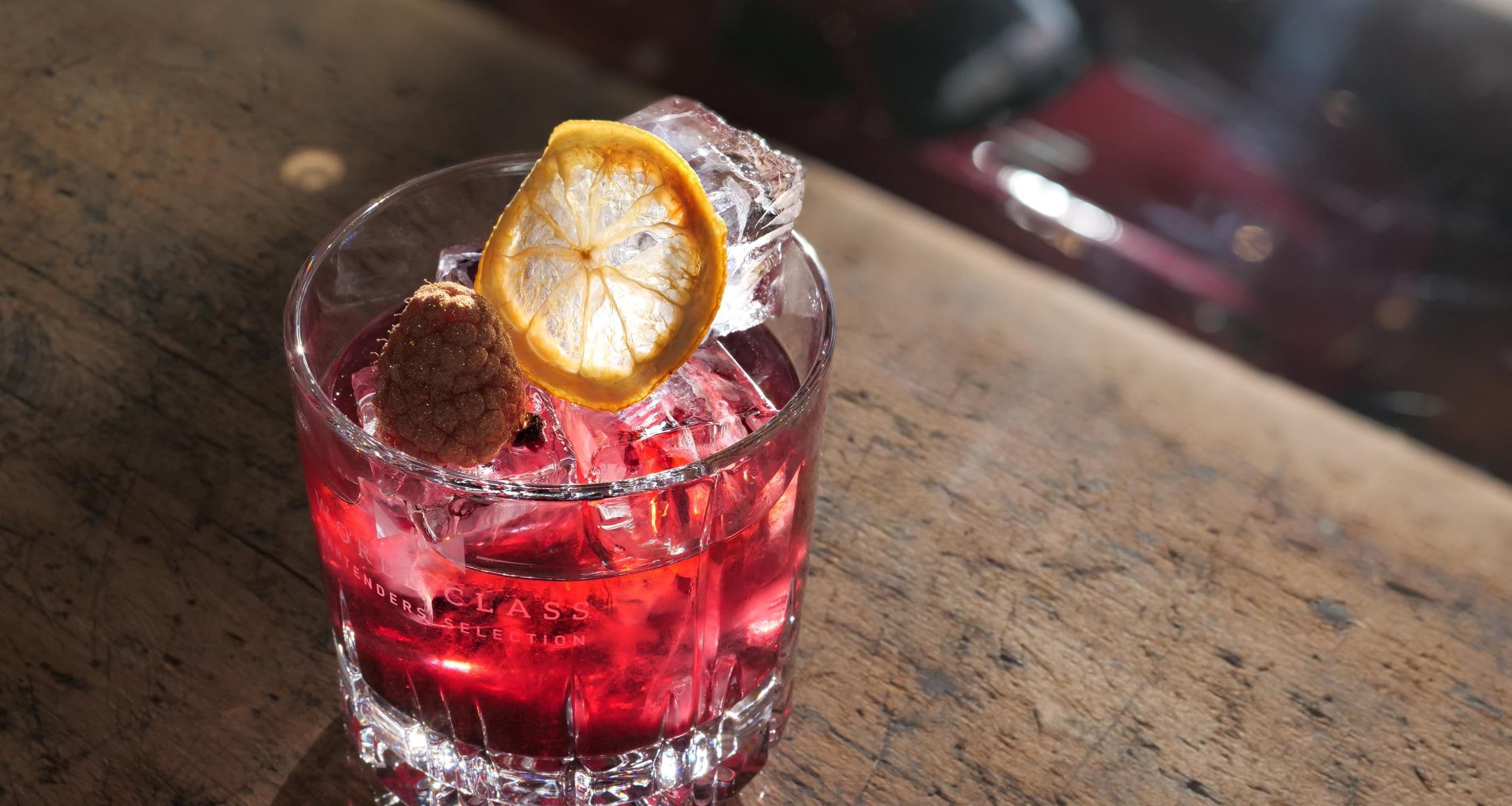 Un vaso con líquido rojo, cubitos de hielo, una rodaja de naranja deshidratada y un objeto marrón, posiblemente una galleta, sobre una superficie de madera.