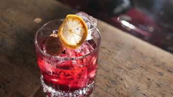 Un vaso con líquido rojo, cubitos de hielo, una rodaja de naranja deshidratada y un objeto marrón, posiblemente una galleta, sobre una superficie de madera.