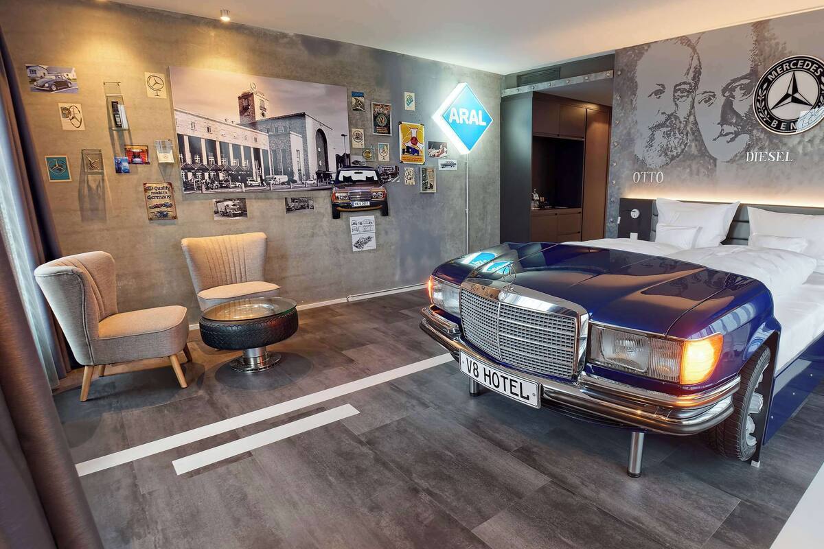 Una habitación de hotel con un coche clásico como cama, un gran mural de Mercedes-Benz y una placa de matrícula que dice 'V8 Hotel'.