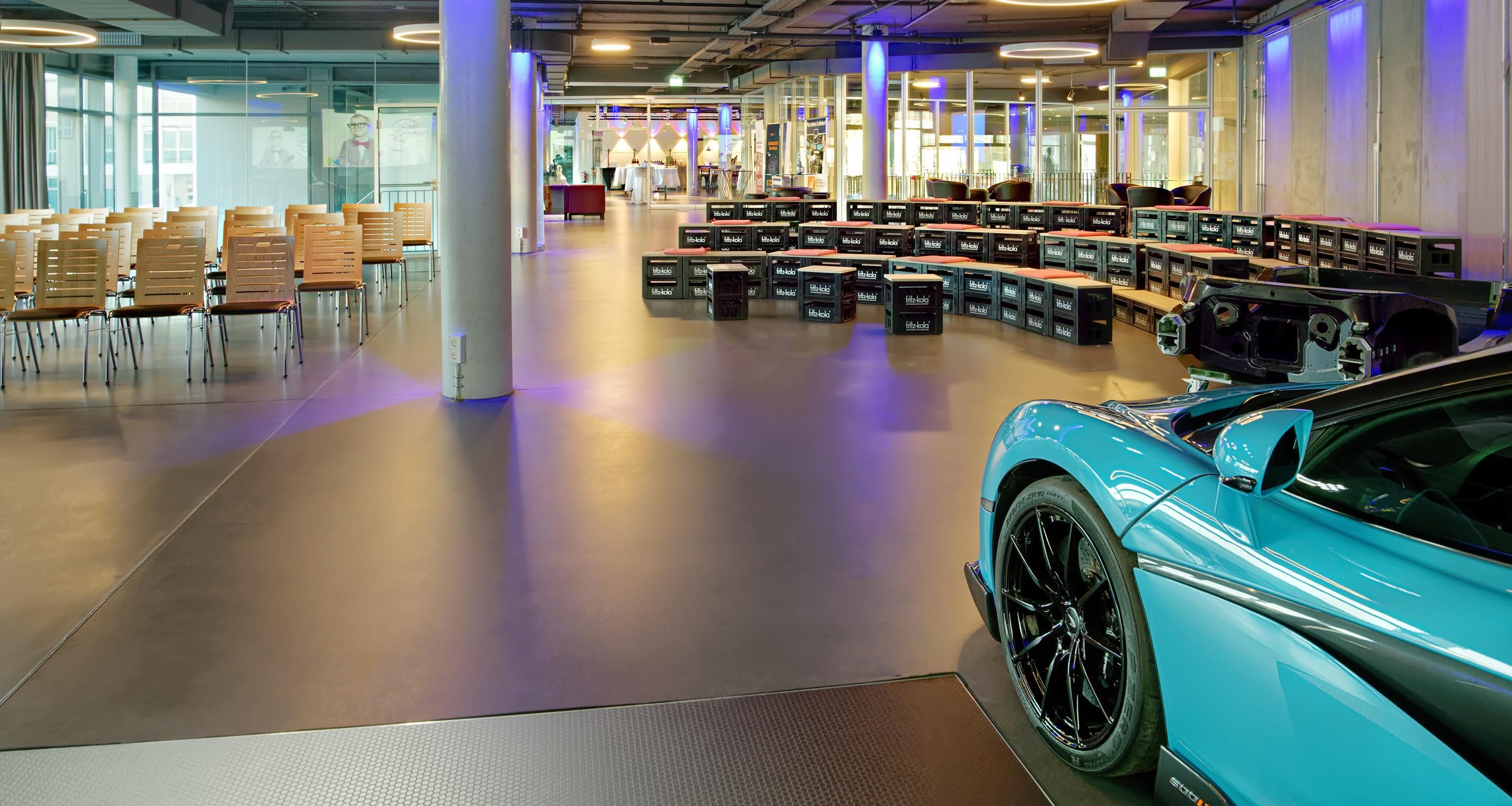 Un vaste espace intérieur avec des rangées de chaises et de tables, une voiture de sport bleue et une grande colonne.