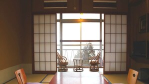 A tatami mat room with a low table and three chairs, large windows with shoji screens, and a view of trees outside.