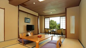 A traditional Japanese room with tatami flooring, a wooden table, purple cushions, a television, and a view of greenery through large windows.