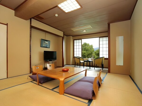 Una habitación japonesa tradicional con suelo de tatami, una mesa de madera, cojines morados, un televisor y vistas a zonas verdes a través de amplios ventanales.