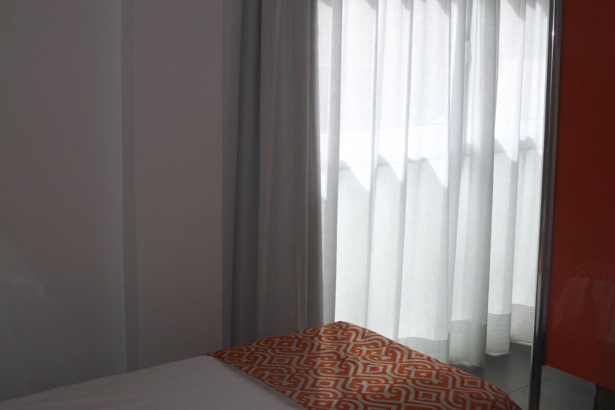 A hotel room with a bed, a window with sheer curtains, and a partial view of a red wall.
