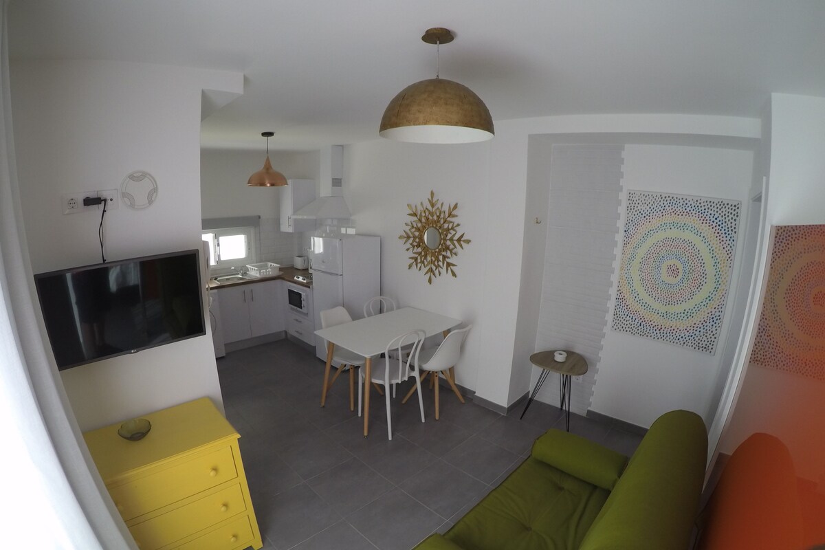A modern kitchen with white cabinets, a dining table with chairs, a large flat-screen TV, and a yellow dresser.