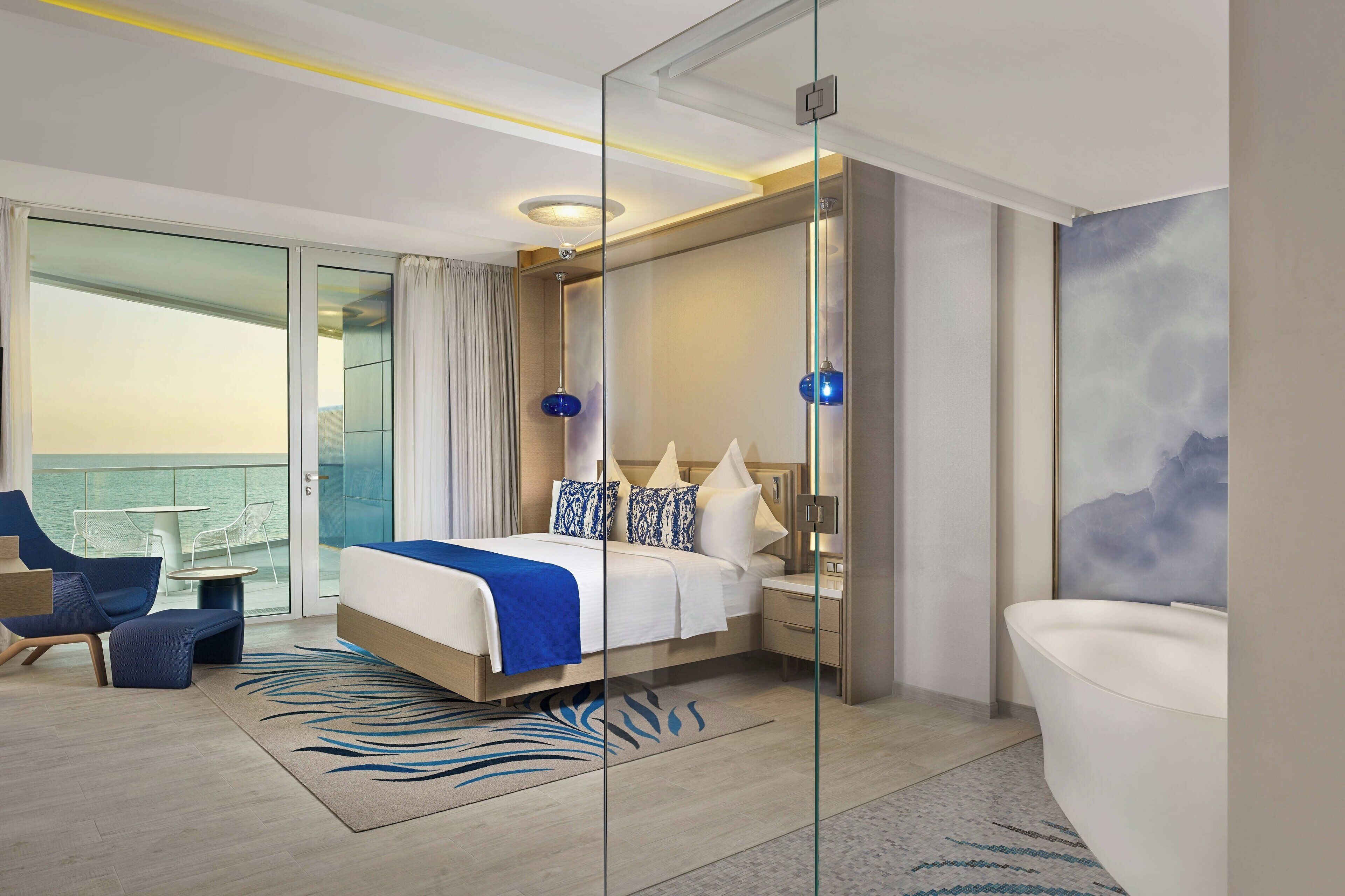 Une chambre d’hôtel moderne dotée d’un grand lit, d’un balcon fermé par des baies vitrées, d’un coin salon avec un fauteuil et une table, et d’une salle de bain avec baignoire.