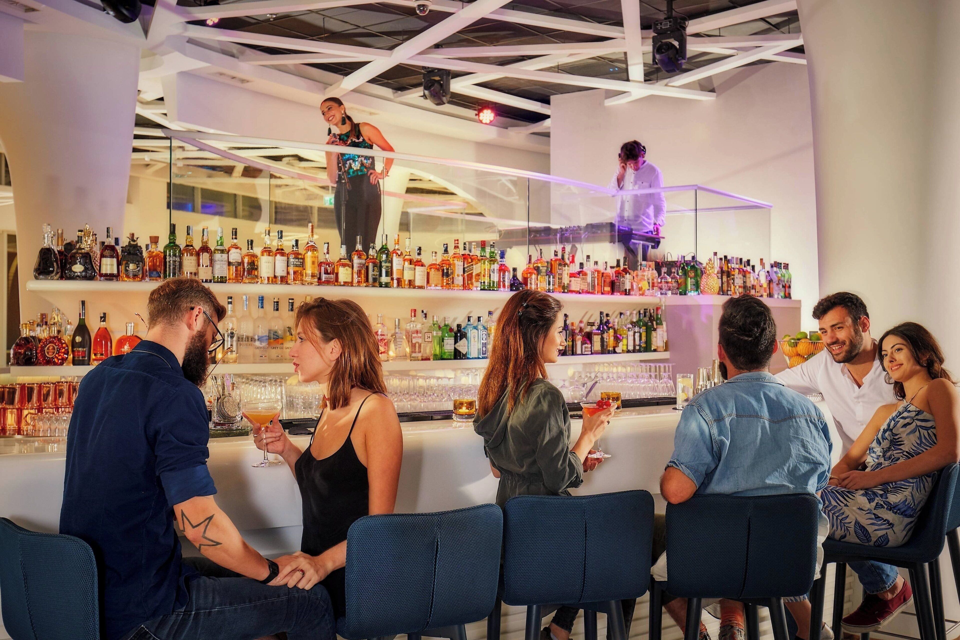 Zona de bar con varios clientes sentados en mesas, un cantinero preparando bebidas y una variedad de botellas y vasos en estantes.