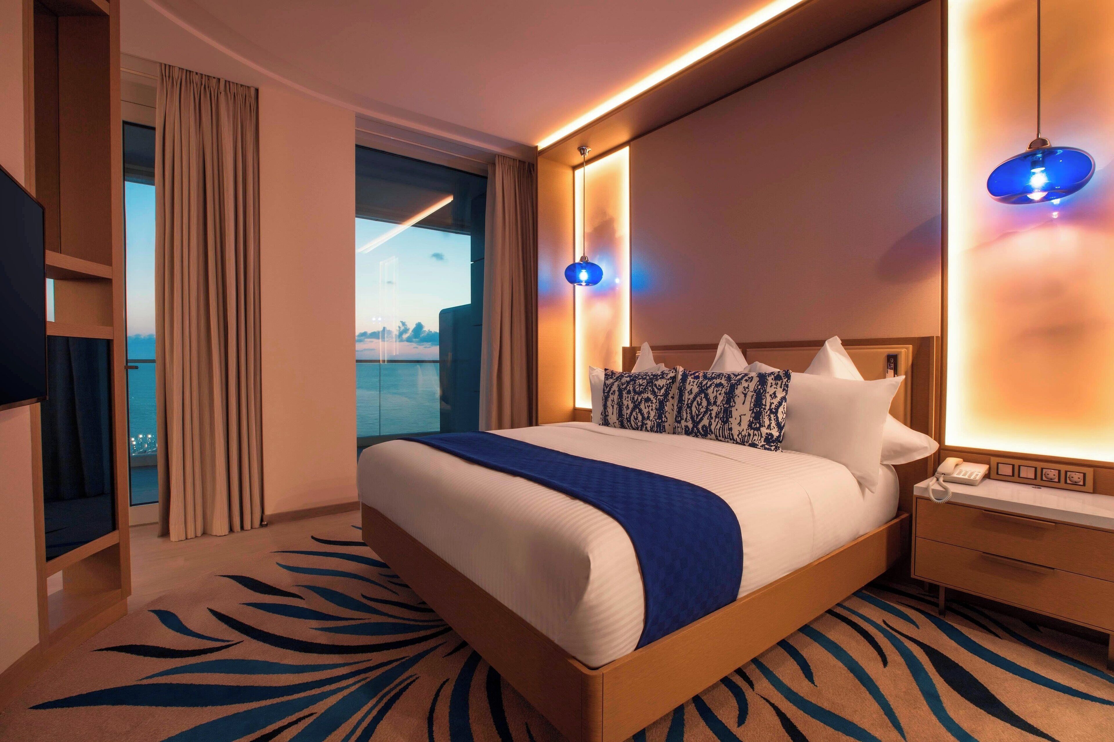 A hotel room with a large bed, a bedside table with a lamp, a view of the city through the window, and a wooden nightstand.
