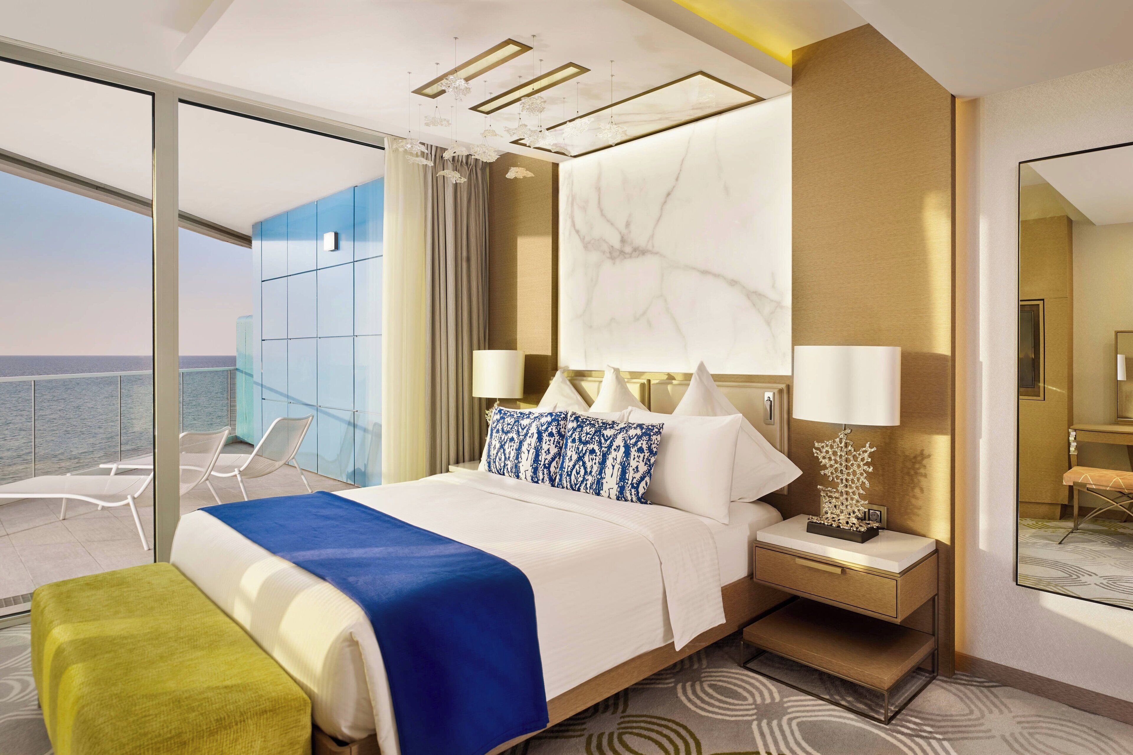 A modern hotel room with a large bed, a nightstand, a lamp, and a view of the sea through floor-to-ceiling windows.