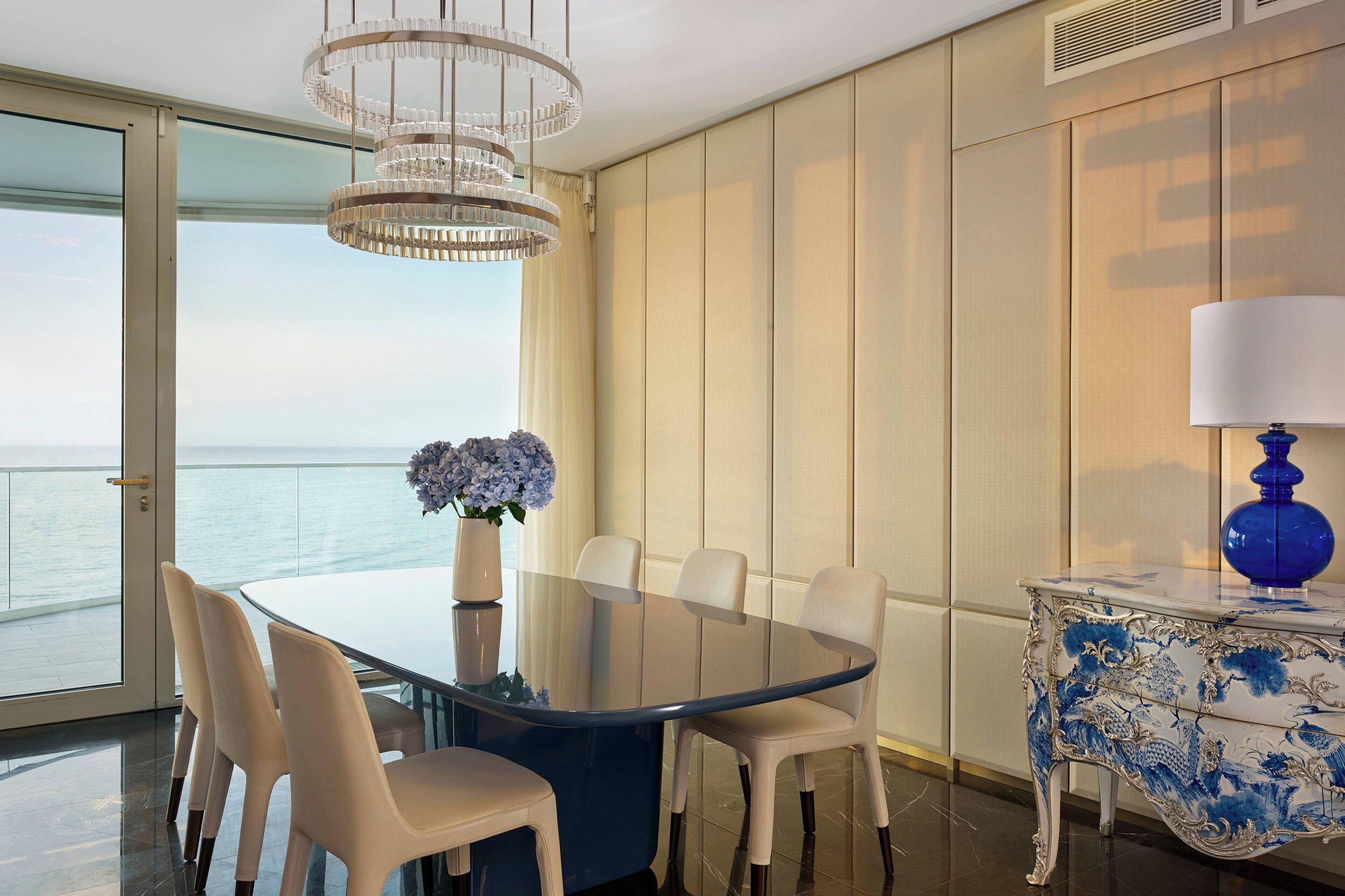 A modern dining area with a glass table, white chairs, and a view of the sea.