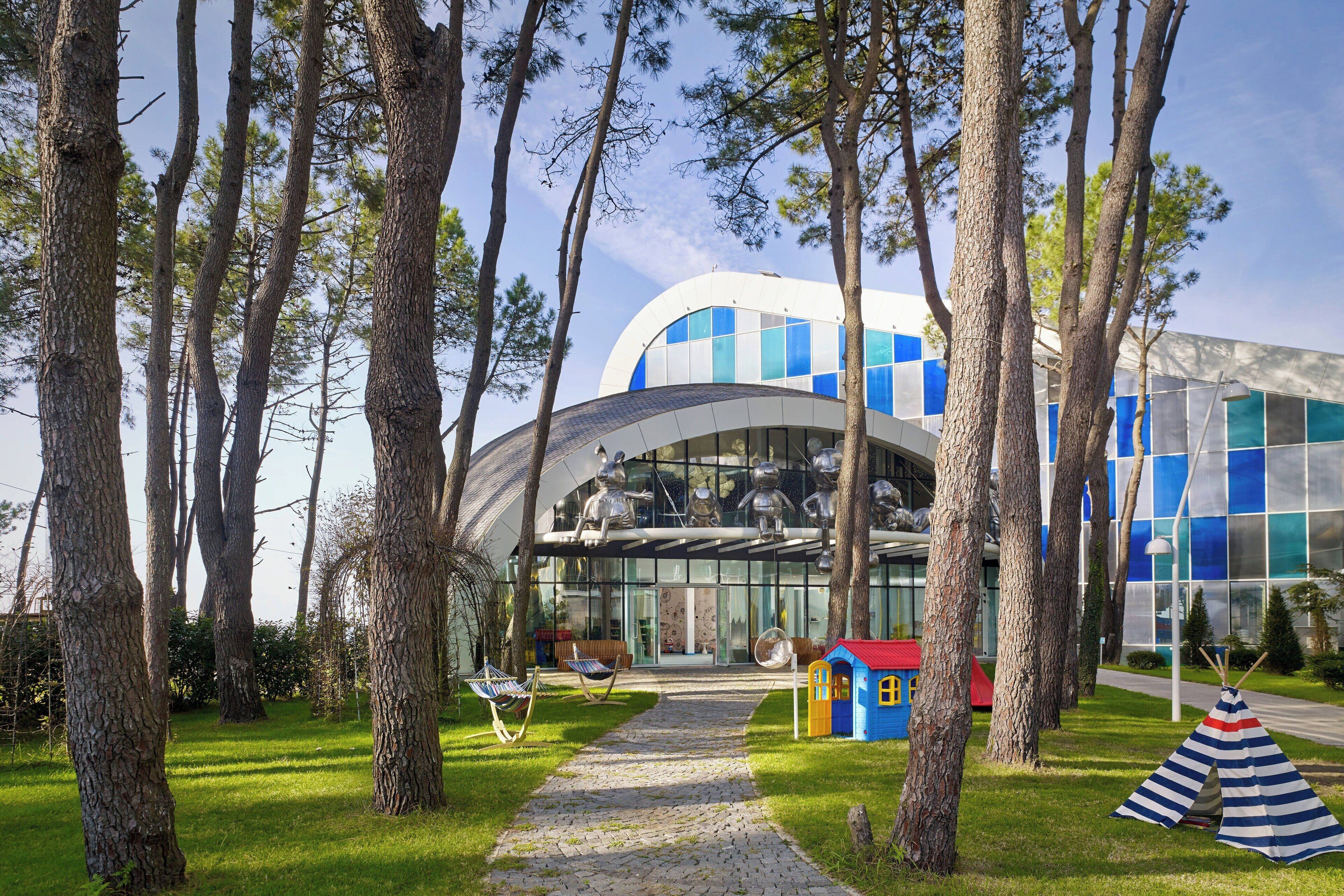 Un bâtiment moderne à la façade incurvée en verre, entouré de grands arbres et d’une zone herbeuse avec un sentier menant à l’entrée.
