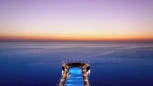 Una piscina larga y estrecha con sillas de descanso y sombrillas, rodeada por una terraza con luces, con vistas a un mar tranquilo al atardecer.