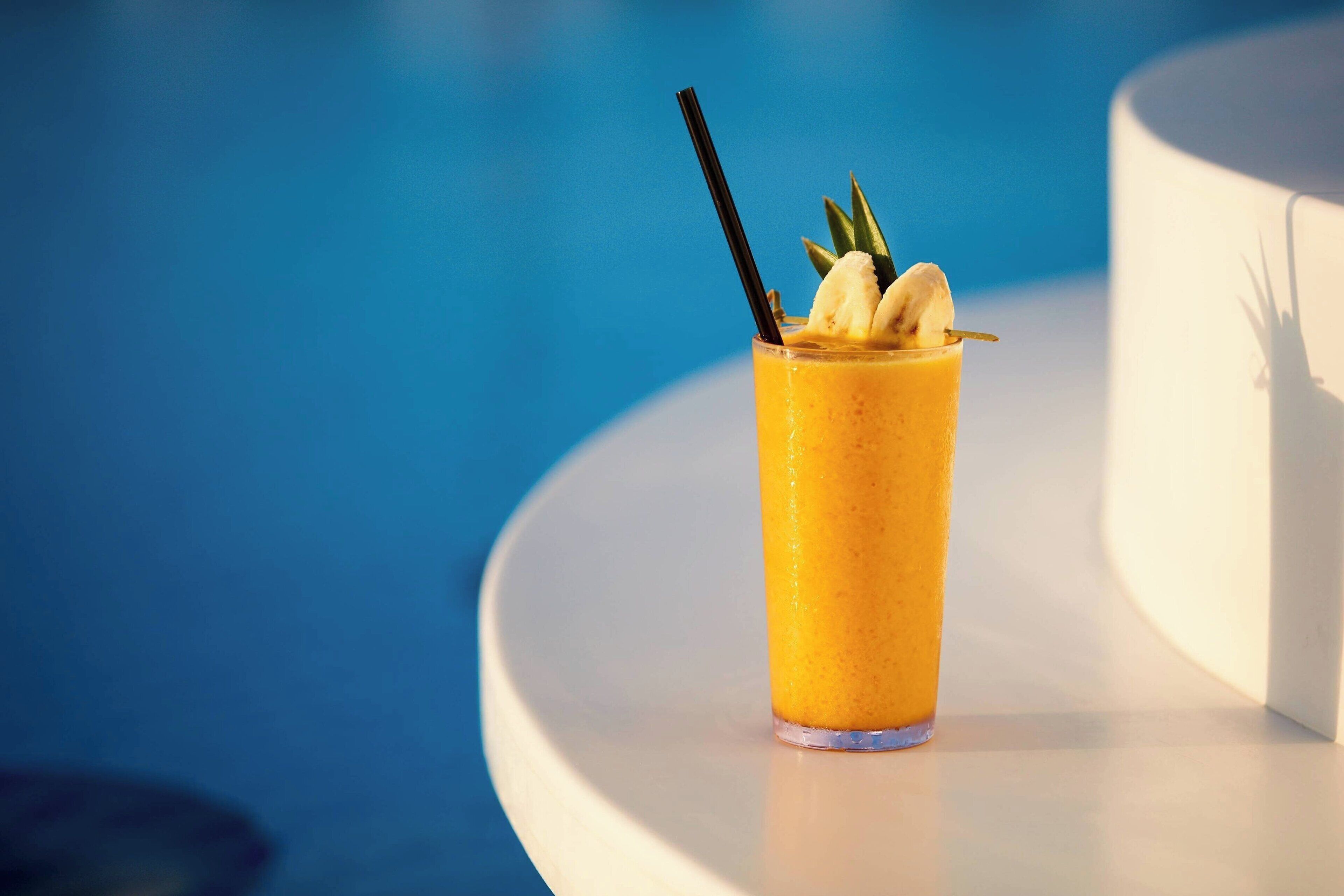 A tall glass of orange smoothie with a straw and a banana slice on a white table near a pool.