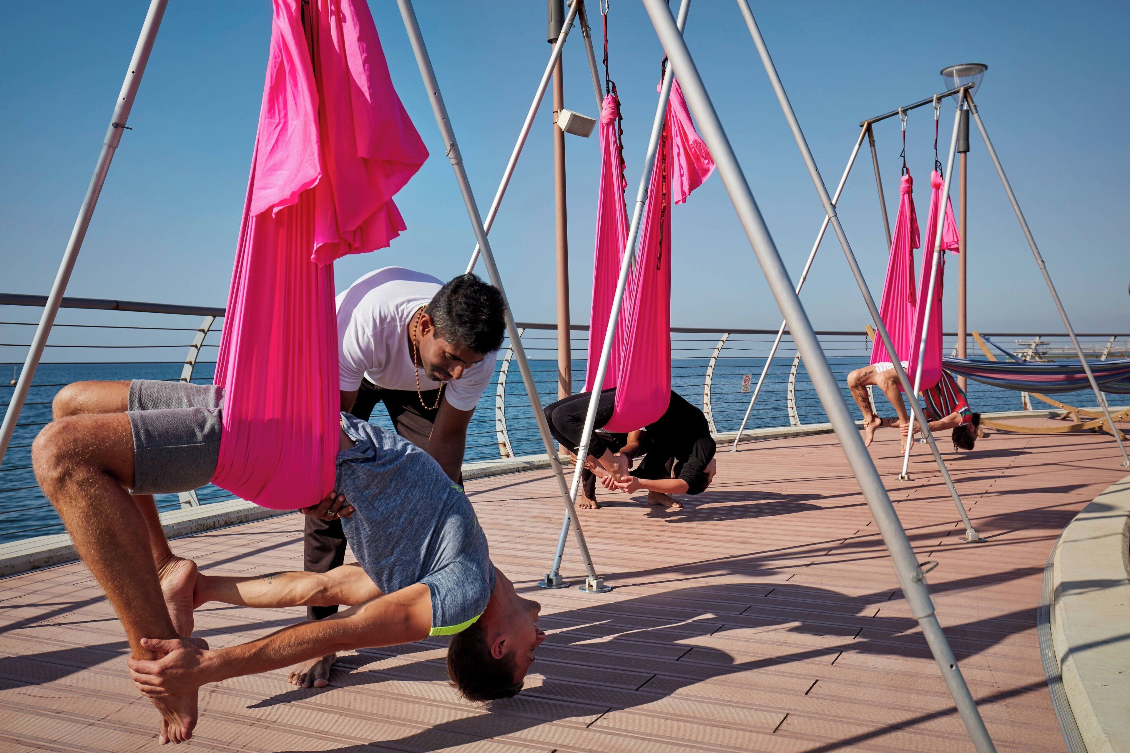 A group of people performing aerial yoga on a deck with pink hammocks, overlooking a body of water.