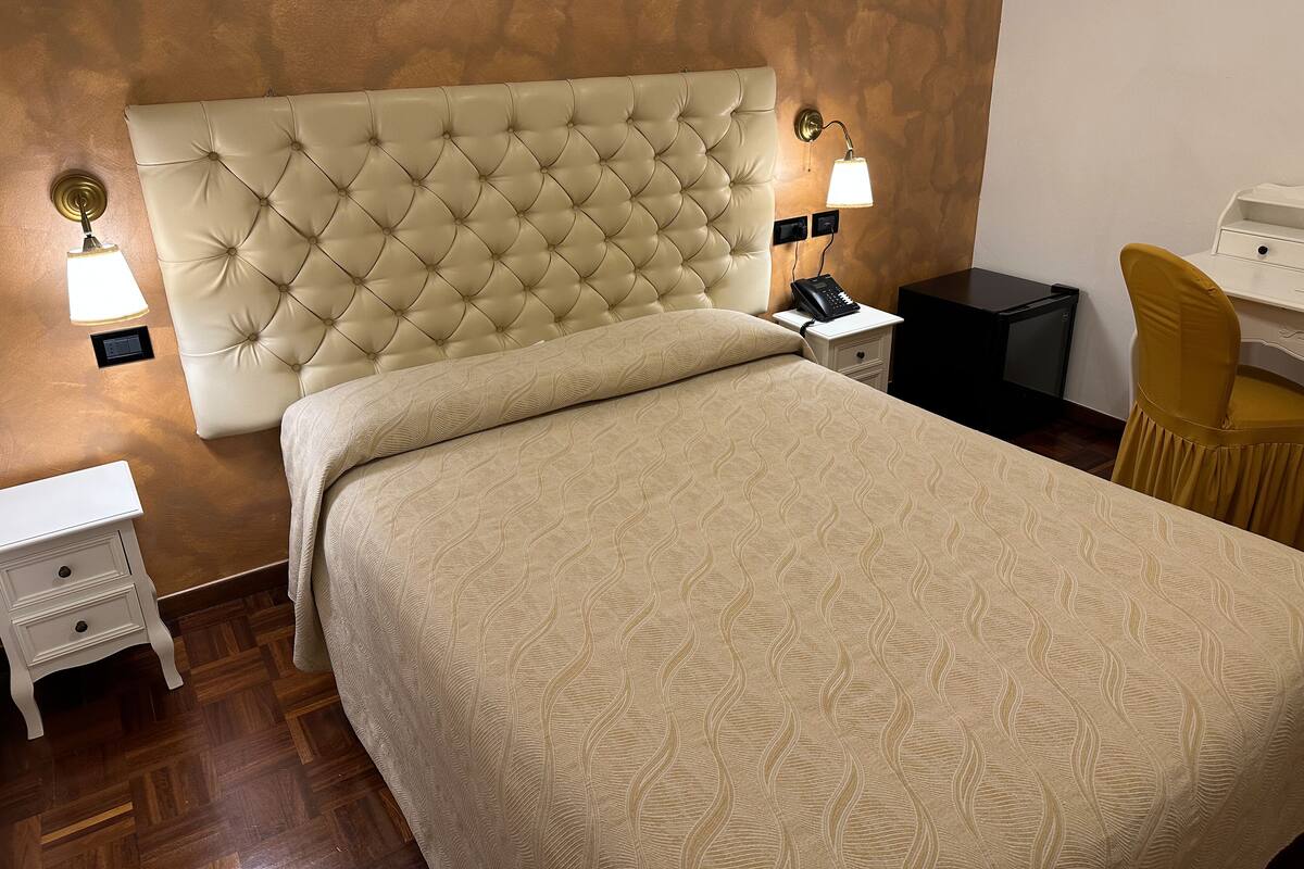 Una habitación de hotel con cabecera acolchada, cama con colcha estampada, lámparas de noche, mesita de noche, escritorio y una silla.