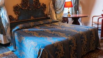 Un dormitorio con una cama grande, un cabecero decorado, cortinas estampadas, una mesita de noche con lámpara y una silla.