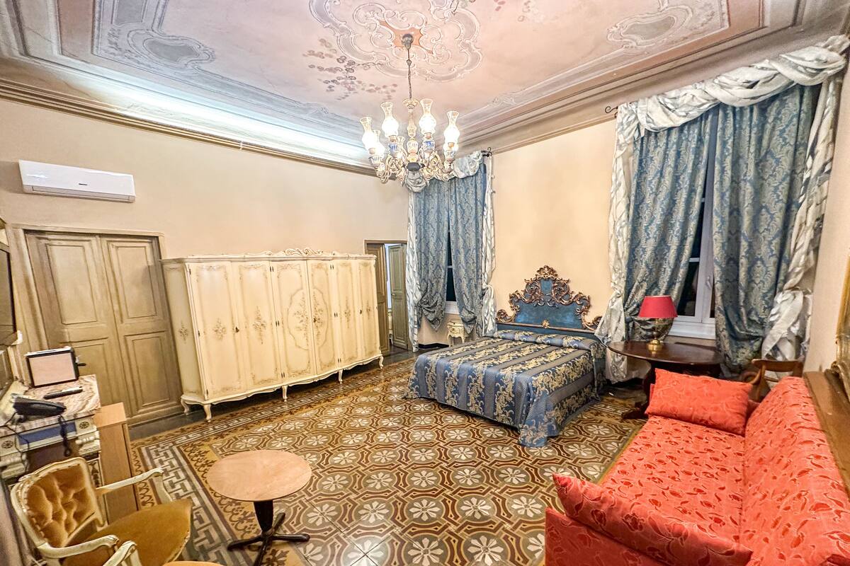 Una habitación con piso estampado, una cama con cabecera azul, un sofá rojo, una araña y un aparador.