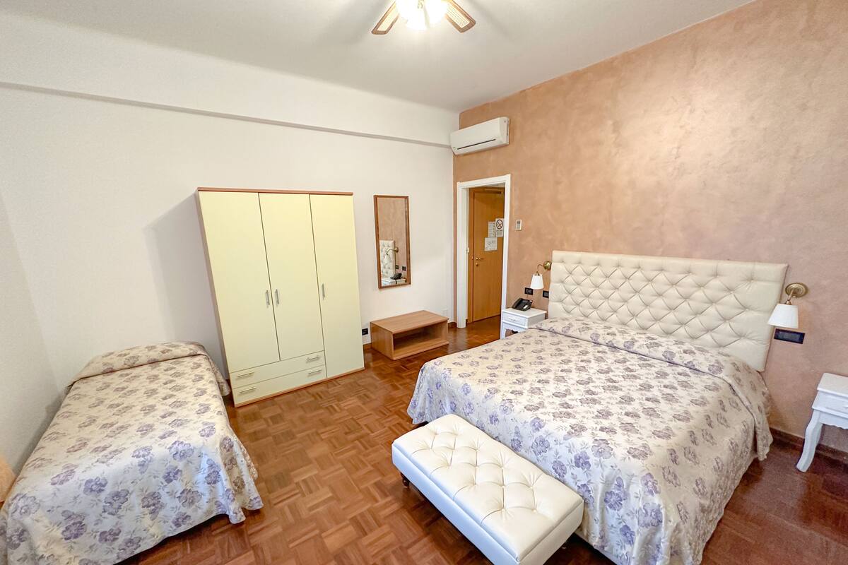 Un dormitorio con una cama grande, una cama más pequeña, un armario, un banco, una mesita de noche y un equipo de aire acondicionado.