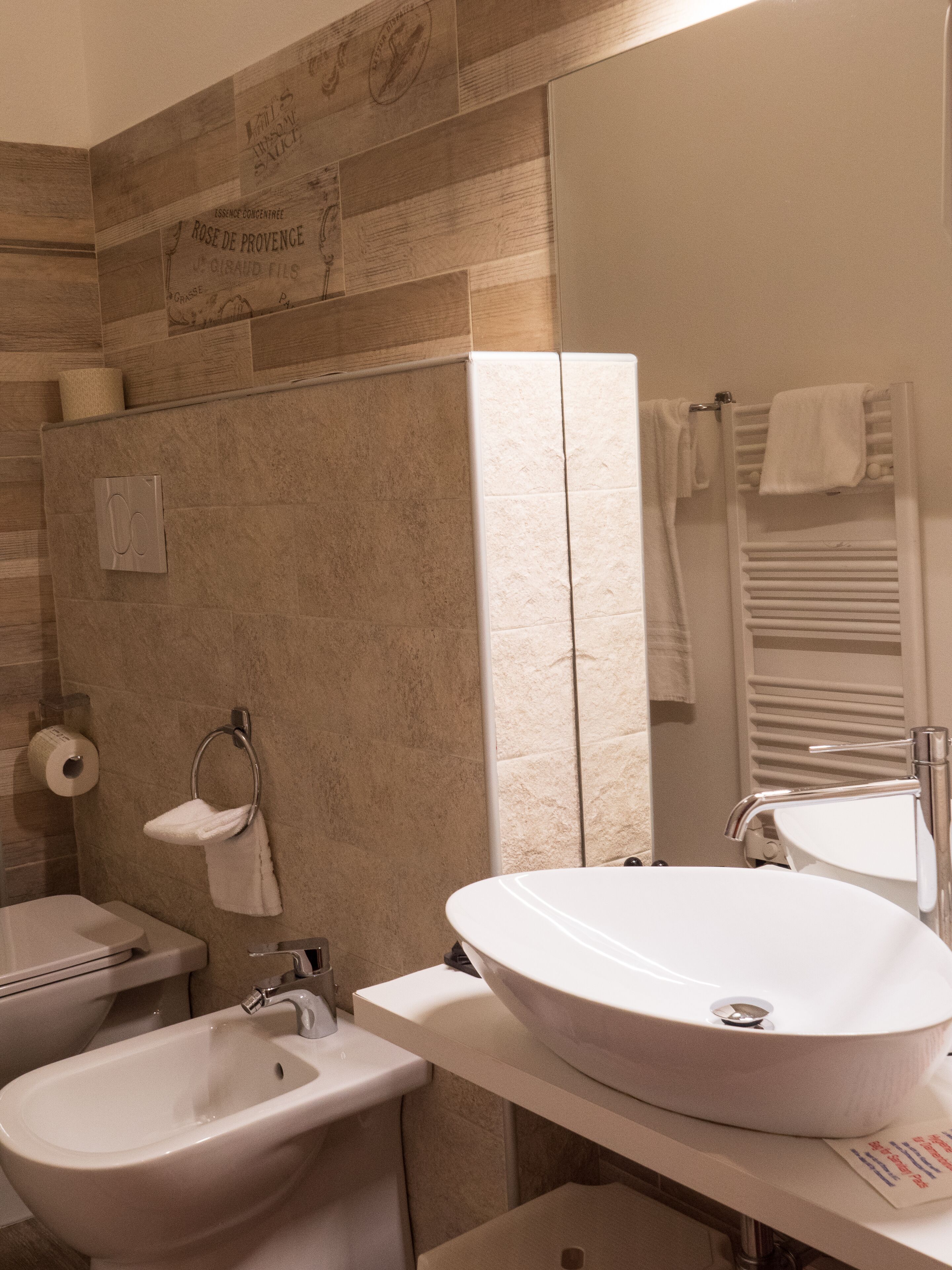 A modern bathroom with a beige tiled wall, a large white sink, a bidet, and a towel rack.