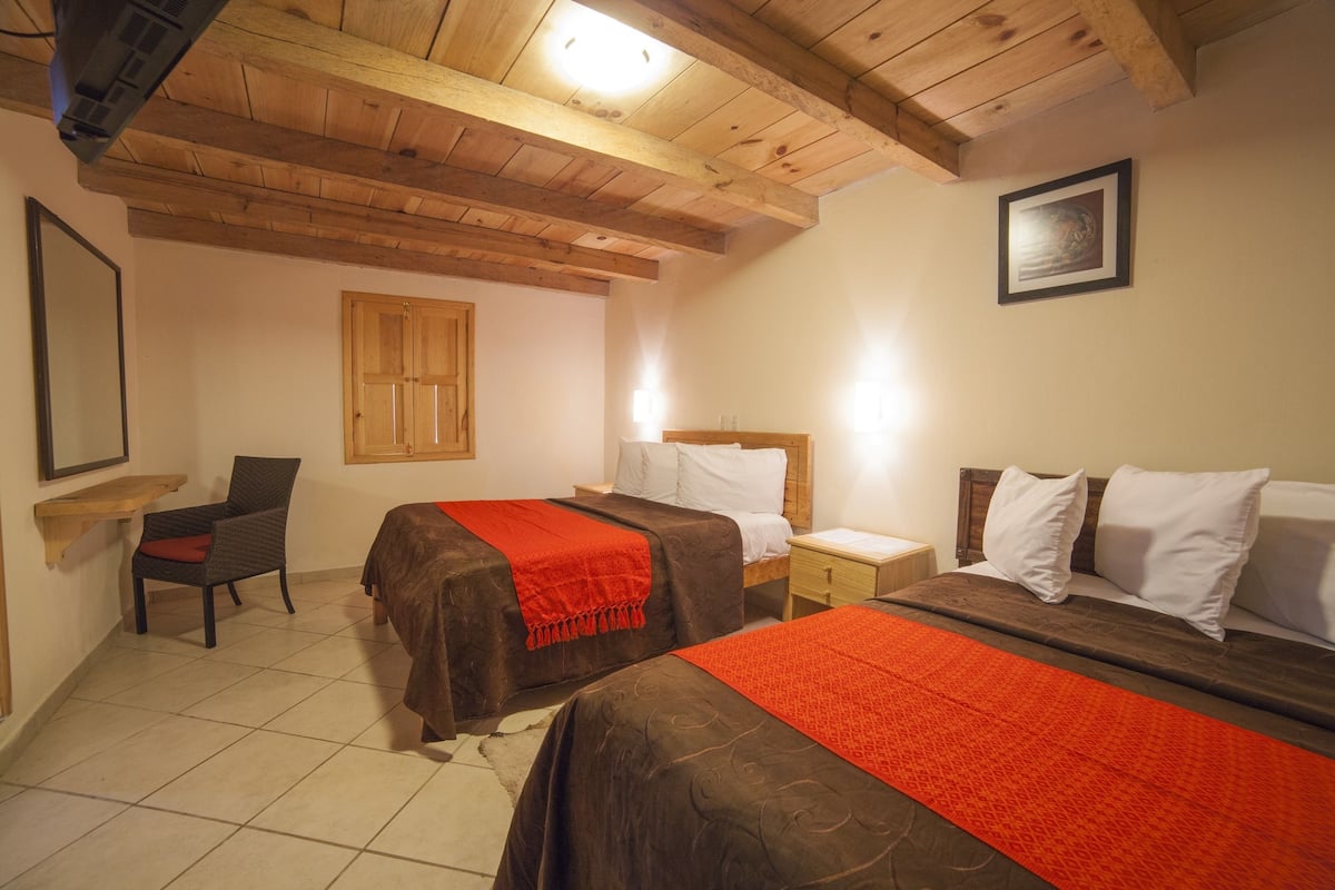 Un dormitorio con dos camas, vigas de techo de madera, una silla, una mesita de noche y un cuadro enmarcado en la pared.