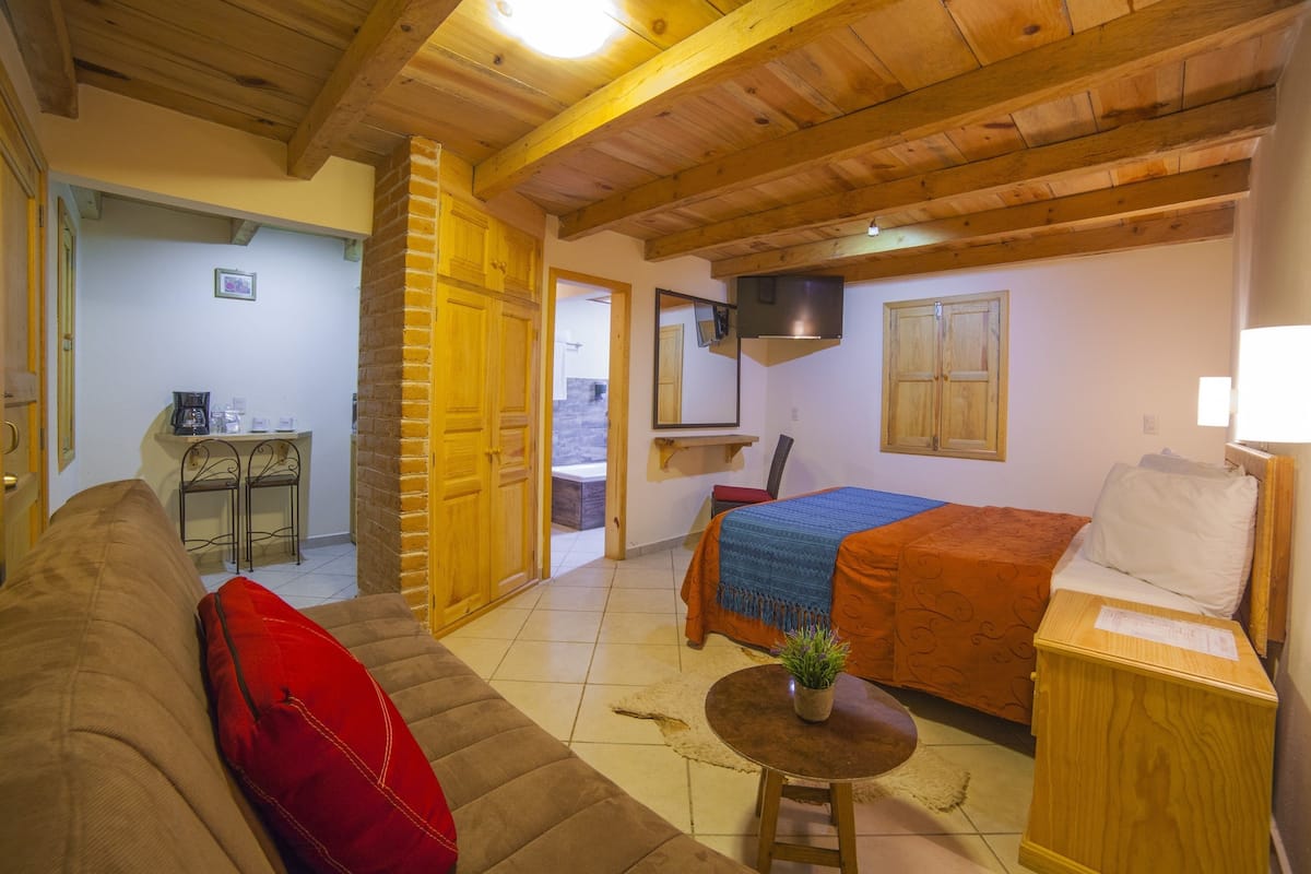 Una habitación acogedora con techo de madera, una cama con manta naranja, un cojín rojo sobre un sofá marrón y una mesita con una planta.