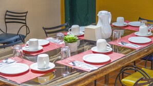 Una mesa de comedor dispuesta con platos blancos, tazas y una jarra, rodeada de sillas.