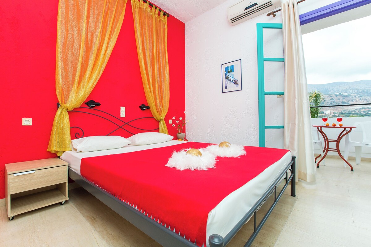 Quarto com cama vermelha, cortinas amarelas, mesa de cabeceira branca e vista para o exterior.