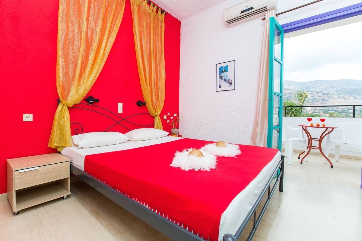Una habitación con cama roja, una mesita de noche pequeña, una mesita auxiliar blanca y un balcón con mesa y sillas.