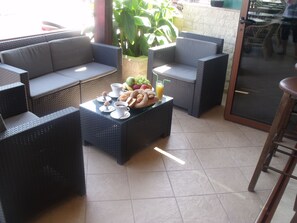 Un patio con mobiliario de mimbre, una mesa con alimentos para el desayuno y una planta en maceta.