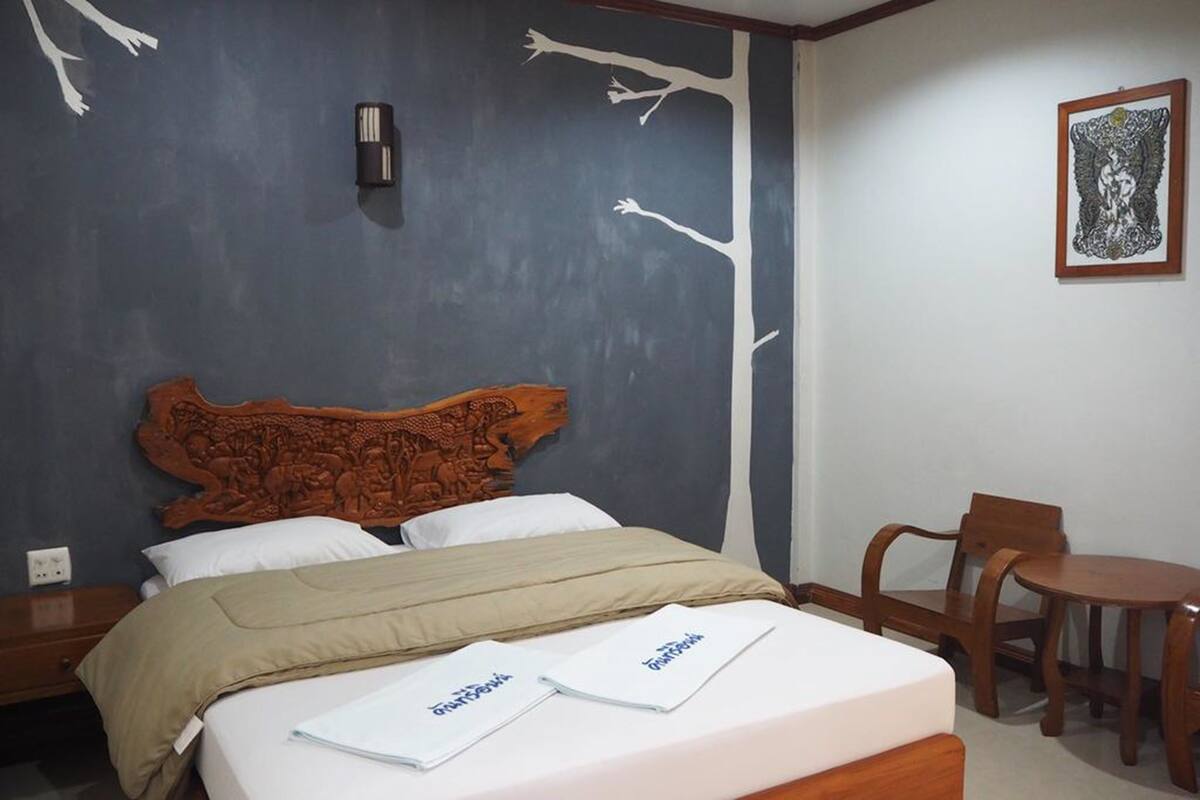 A bedroom with a bed, a nightstand, a chair, a small table, and a wall with a tree design.