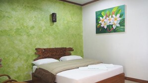 A bedroom with a wooden bed, green textured walls, a painting of flowers, and a small wooden table. - Country Inn 304 (Si Maha Phot)