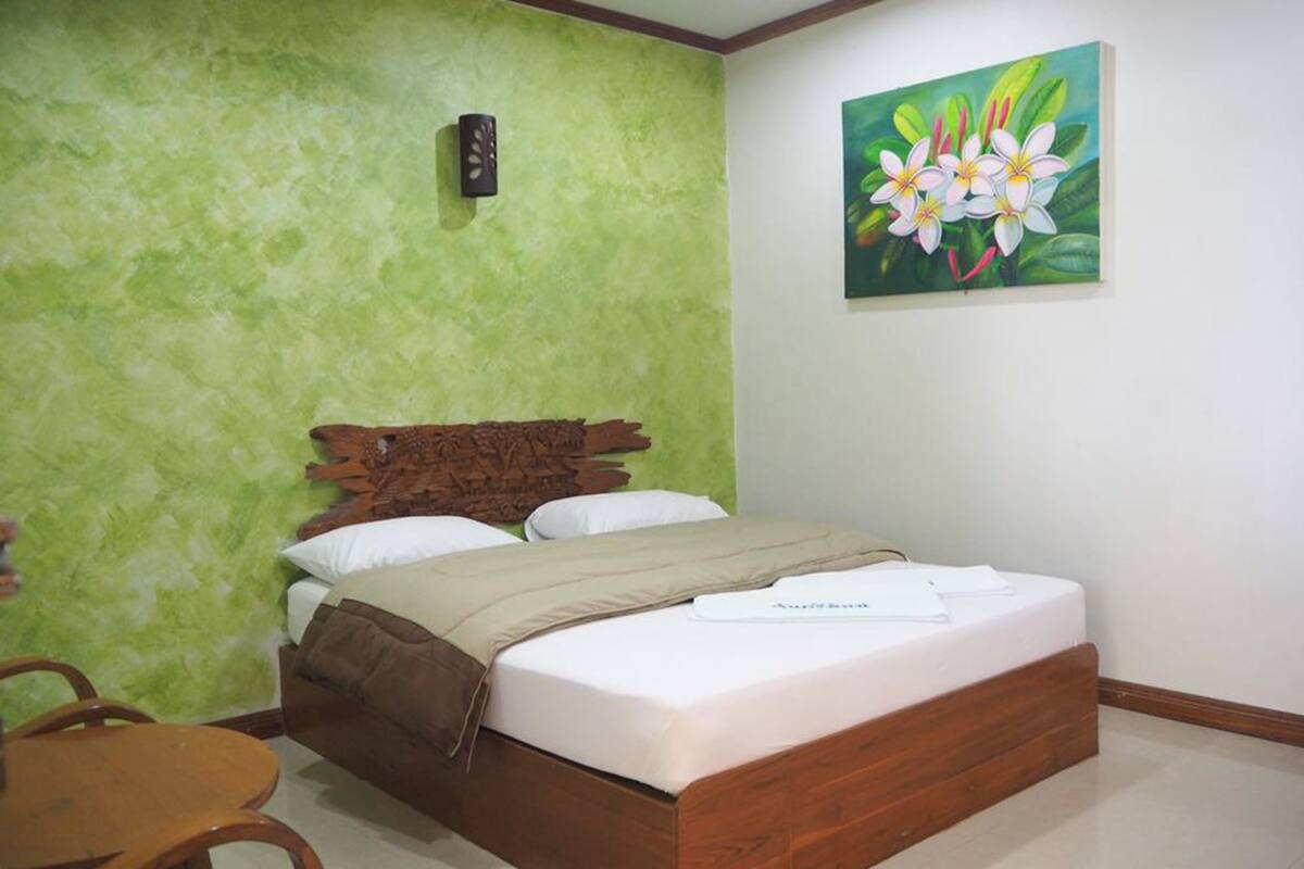 A bedroom with a wooden bed, green textured walls, a painting of flowers, and a small wooden table.