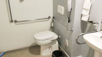 A bathroom with a toilet, a sink, a handrail, a phone, and a trash bin.