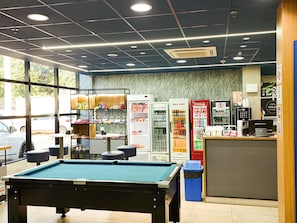 Een biljart, automaten en een balie met een computer in een ruimte met een betegelde vloer en een raam.