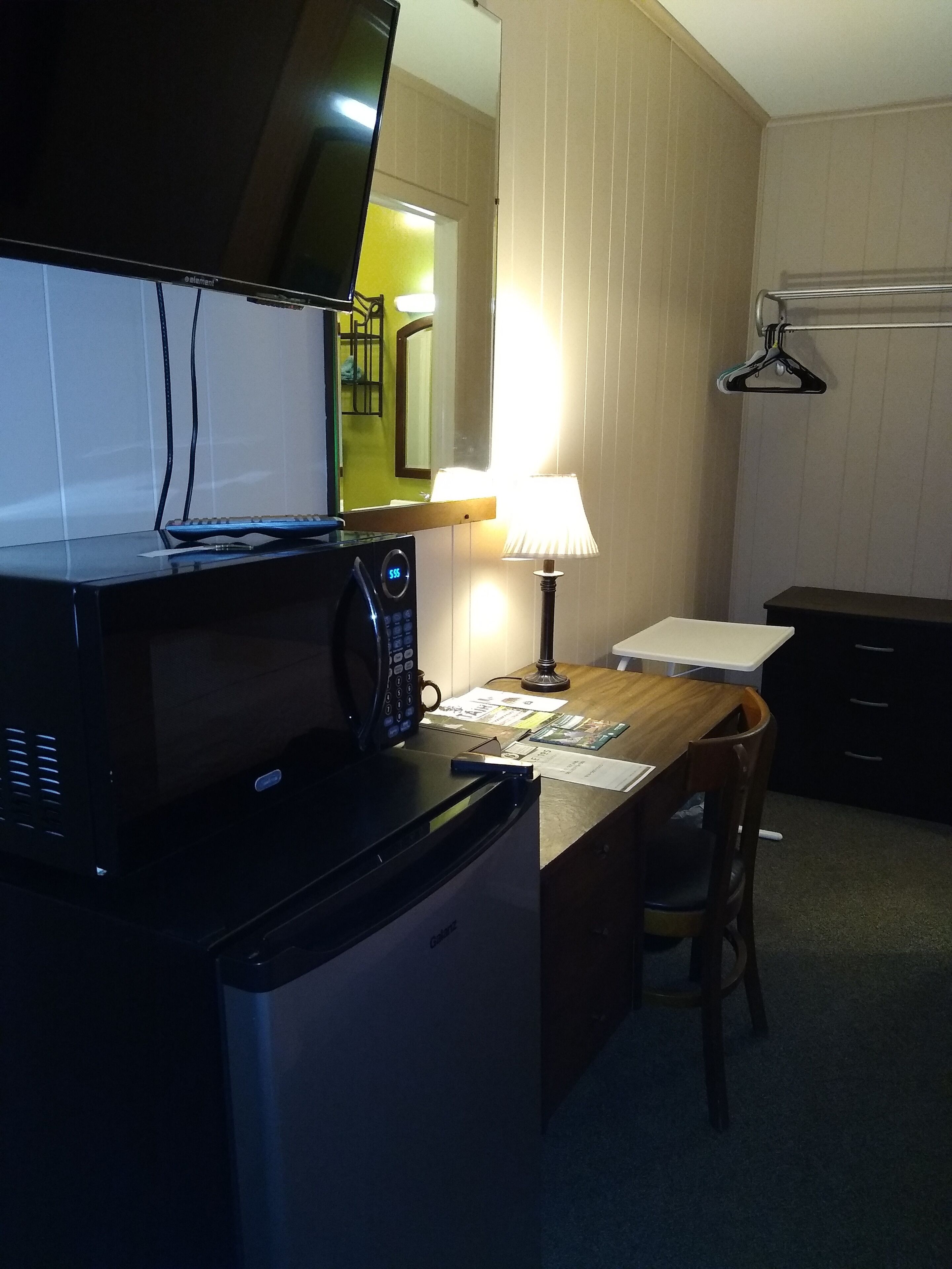 A compact kitchen with a microwave, a small refrigerator, a wooden table, and a chair. A lamp provides light, and a mirror hangs above the counter.
