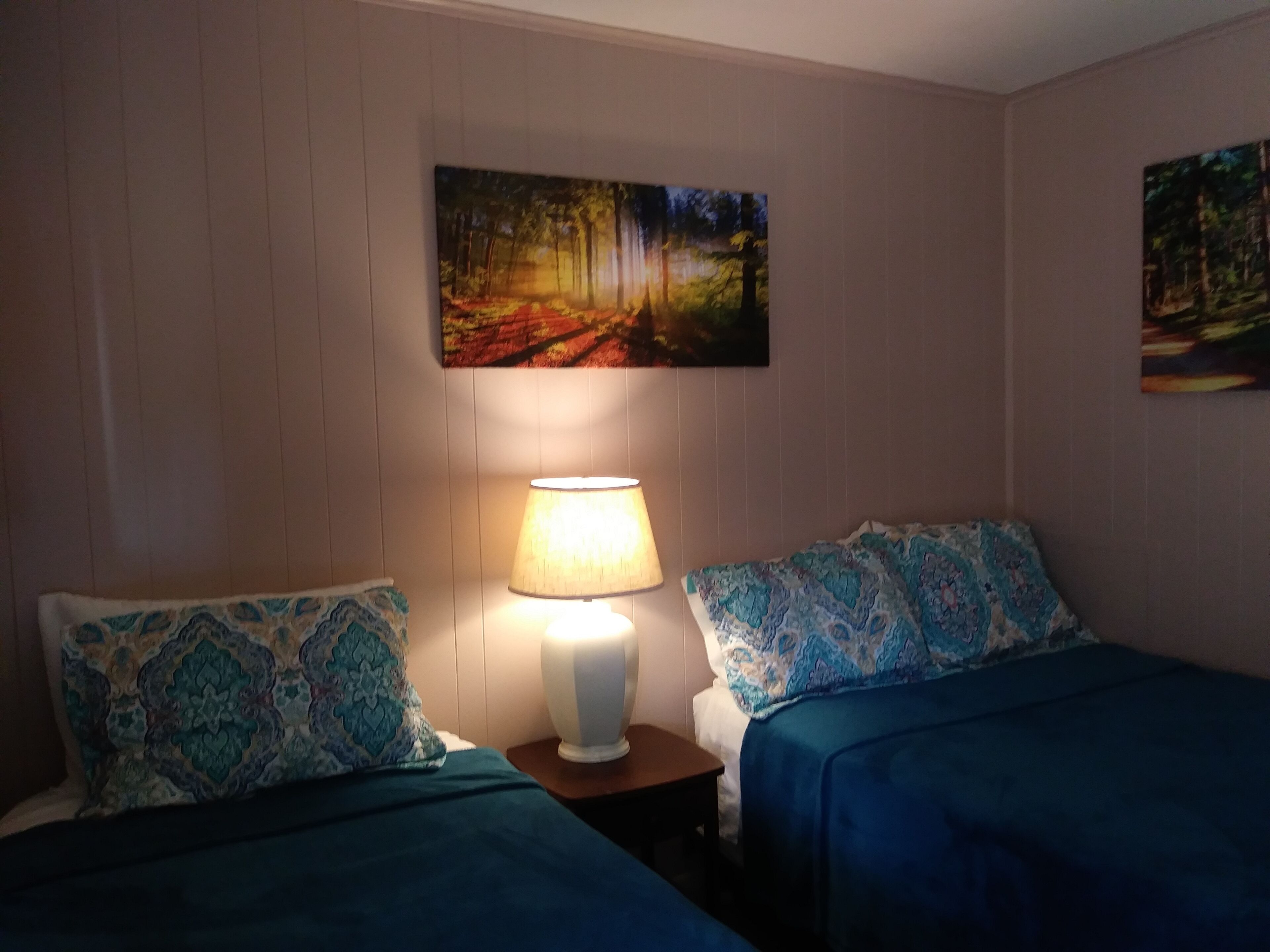 Quarto com duas camas, uma mesa de cabeceira com abajur e duas pinturas nas paredes.