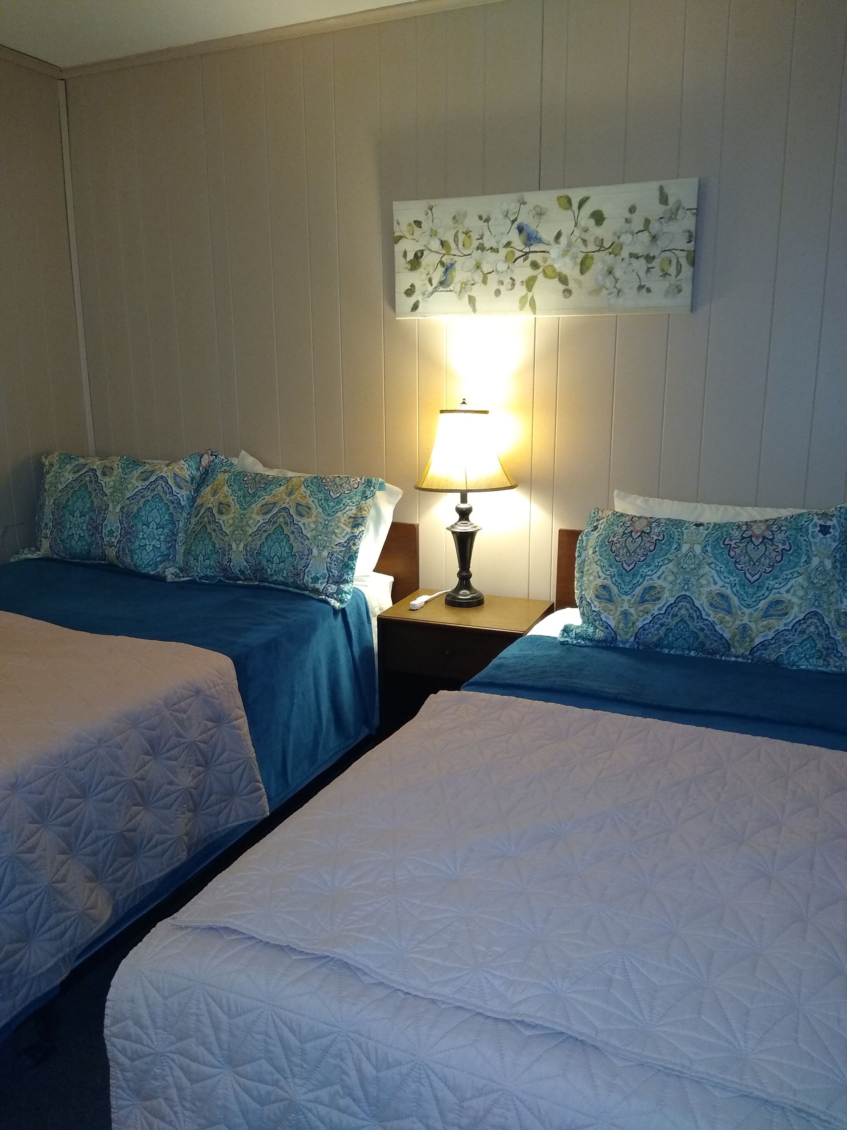 Habitación de hotel con dos camas, una mesita de noche con lámpara y un cuadro floral en la pared.